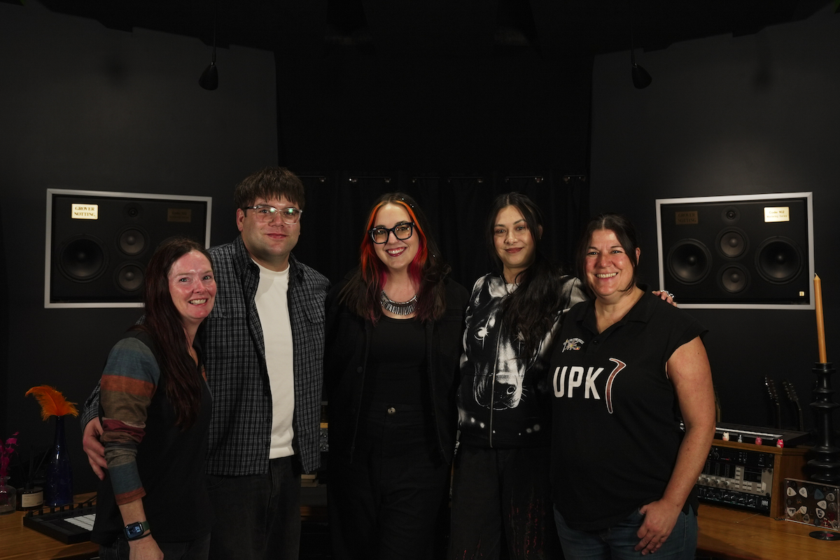 Members of the RISER collective posing together inside a music recording studio with wall-mounted studio monitors in the background.